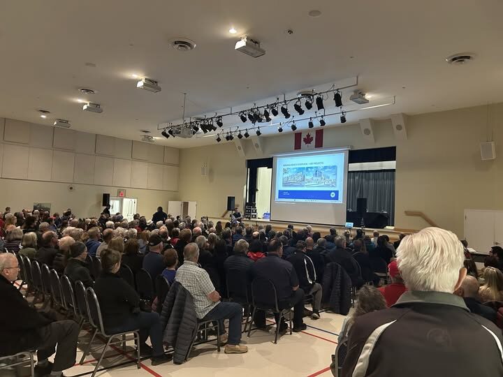 A standing-room-only crowd at a Wasaga Beach community hall meeting, with attendees seated facing a projection screen and a Canadian flag.